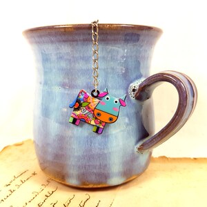 Könnte beinhalten: Eine blaue Keramiktasse mit braunem Henkel und einem bunten Kuh-Tee-Infuser, der am Rand hängt.