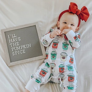 May include: A baby wearing a white onesie with colorful coffee cup designs and a red bow headband. The onesie features various coffee flavors. A gray framed letter board next to the baby reads, "I'LL HAVE THE PUMPKIN SPICE."