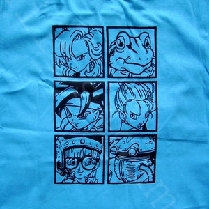 May include: A light blue t-shirt with a black graphic print of six cartoon characters in squares. The characters include a woman with long hair, a frog, a man with spiky hair, a woman with long hair, a woman with short hair and glasses, and a robot.