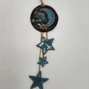 May include: Ceramic wall hanging with a crescent moon and stars. The moon is dark blue with a light blue crescent, set in a brown circular frame. Three blue stars hang below on twine. A decorative celestial theme.
