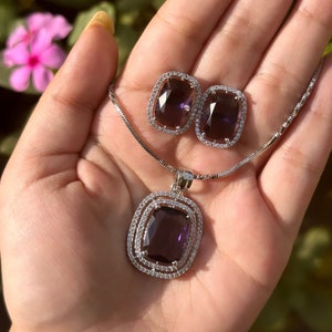 Peut inclure: Un ensemble de bijoux argentés comprenant un collier pendentif et des boucles d'oreilles assorties. L'ensemble comprend une grosse pierre précieuse rectangulaire violet foncé entourée d'un halo de petits cristaux clairs.