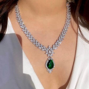 May include: A silver necklace with a teardrop-shaped green gemstone pendant surrounded by smaller clear stones. The necklace is made of a chain of clear stones in a leaf-like pattern.