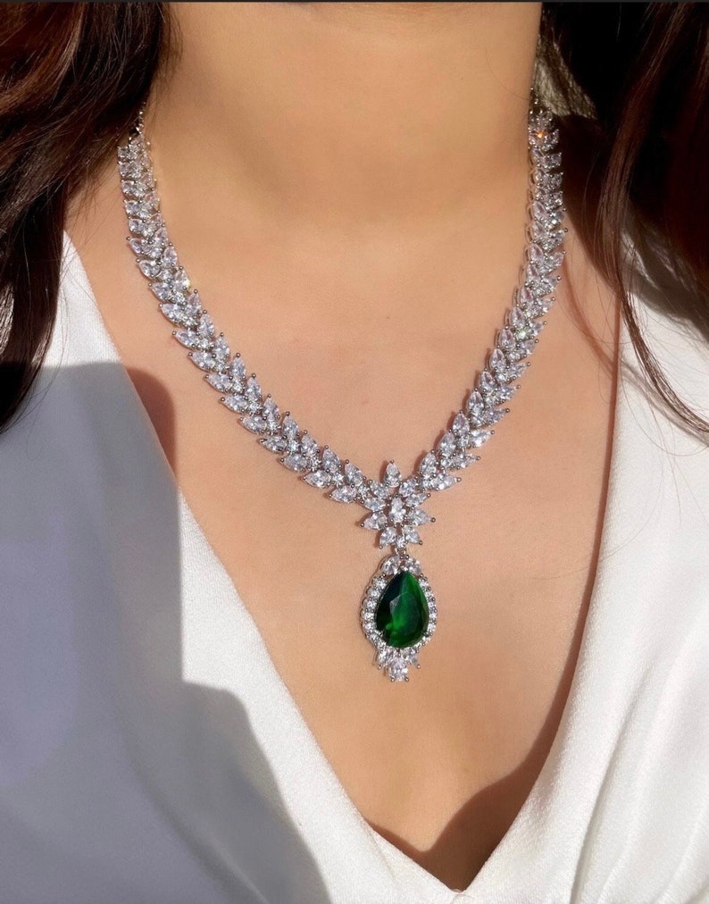 May include: A silver necklace with a teardrop-shaped green gemstone pendant surrounded by smaller clear stones. The necklace is made of a chain of clear stones in a leaf-like pattern.