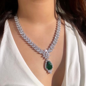 May include: A silver necklace with a large pear-shaped green gemstone pendant surrounded by smaller clear stones. The necklace is made of a chain of clear stones in a leaf-like pattern.