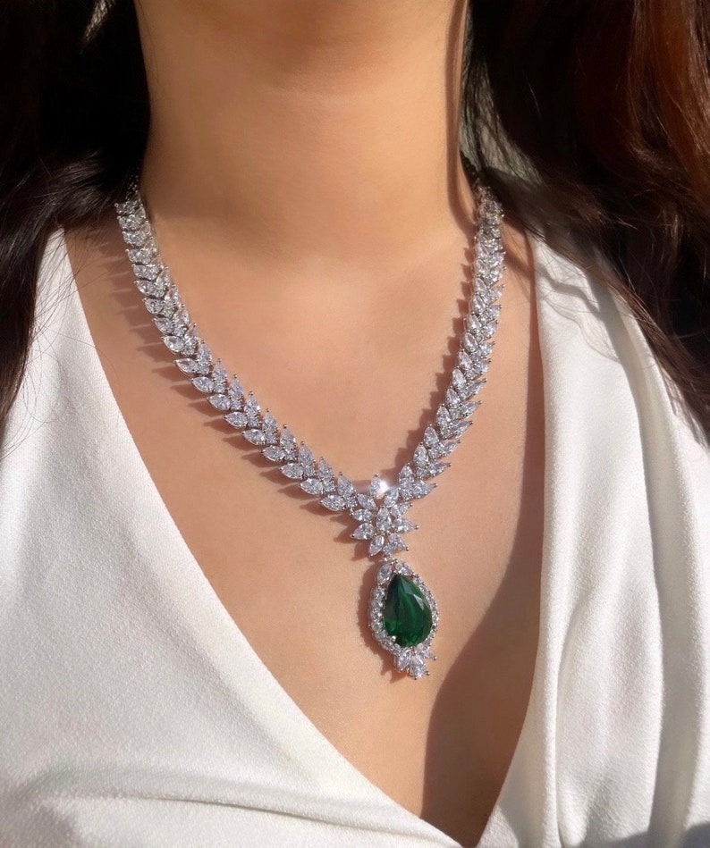 May include: A silver necklace with a large pear-shaped green gemstone pendant surrounded by smaller clear stones. The necklace is made of a chain of clear stones in a leaf-like pattern.