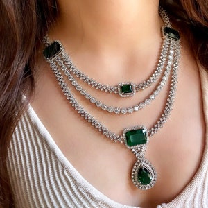 Puede incluir: Un collar de plata con tres hileras de piedras de circonita cúbica y un gran colgante verde esmeralda. El colgante tiene forma de lágrima con una piedra cuadrada verde esmeralda en la parte superior.