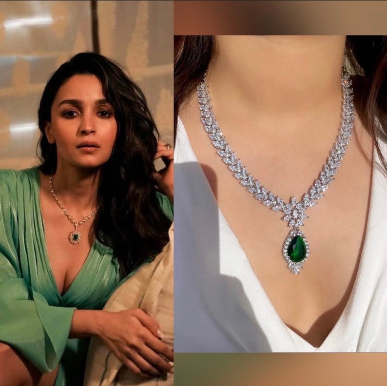 May include: A silver necklace with a large green gemstone pendant. The necklace is made of small, clear stones that are set in a leaf-like pattern. The pendant is a teardrop shape and is surrounded by smaller stones.
