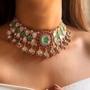 Può includere: Una collana dorata con una grande pietra centrale di smeraldo quadrato, circondata da pietre preziose rosse e trasparenti più piccole. La collana ha un design a pettorina con accenti pendenti a forma di goccia e grappoli di smeraldi verdi e pietre preziose trasparenti.