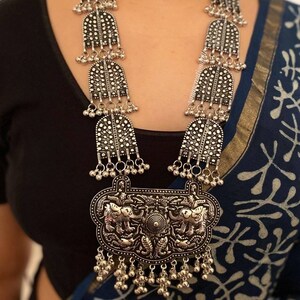 May include: A silver necklace with a large pendant featuring two elephants. The pendant is surrounded by smaller silver beads and bells. The necklace is long and has multiple layers of silver beads and bells.
