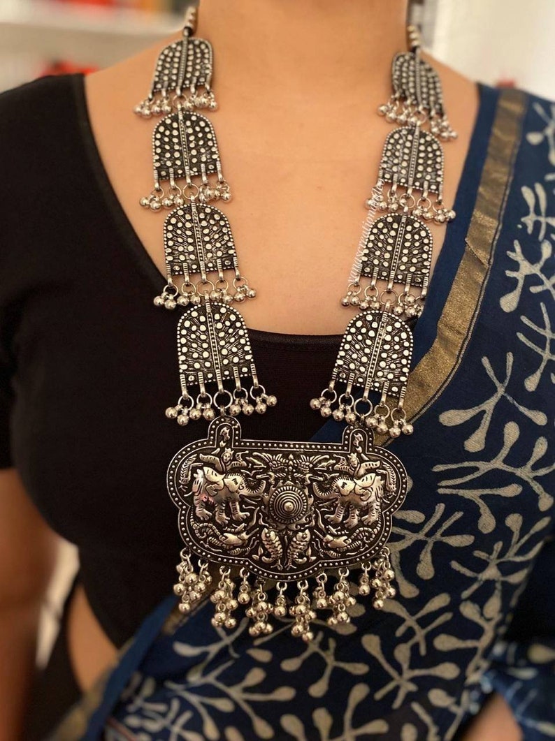 May include: A silver necklace with a large pendant featuring two elephants. The pendant is surrounded by smaller silver beads and bells. The necklace is long and has multiple layers of silver beads and bells.