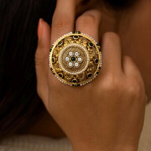 May include: A large, ornate gold ring with a circular design. The ring features a central floral pattern with clear and green gemstones, surrounded by pearls and black accents. The ring is a statement piece of jewelry.
