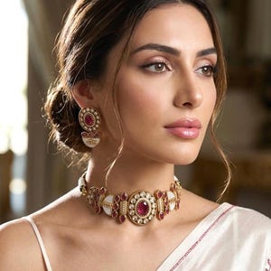 May include: A gold-toned choker necklace and matching stud earrings featuring deep red oval and teardrop-shaped stones, white enamel accents, and small clear crystals. The jewellery set is worn with a white garment featuring subtle red embroidery.