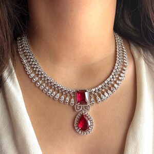 May include: A dazzling necklace featuring a large, square-cut red gemstone pendant with a teardrop-shaped stone below. The necklace is encrusted with numerous small, clear gemstones, creating a sparkling, elegant design. The necklace is worn around the neck.
