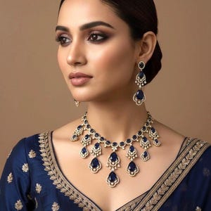 May include: A woman wearing a matching necklace and earring set. The jewelry features large, teardrop-shaped, dark blue gemstones set in a gold-colored metal. The woman is wearing a dark blue top with gold embroidery.