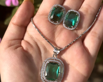 Könnte beinhalten: Ein Schmuckset mit einer Anhängerkette und passenden Ohrringen. Der Anhänger und die Ohrringe haben große, smaragdgrüne, rechteckige Steine, die von einem Kranz aus kleinen, klaren Kristallen umgeben sind. Die Kette und die Ohrringe sind aus silberfarbenem Metall gefertigt.