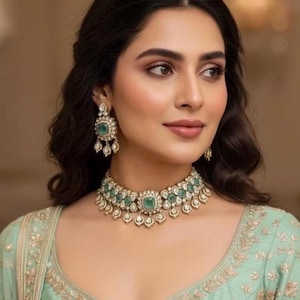 Può includere: Una donna indossa una collana e orecchini dorati con accenti di pietre preziose verde smeraldo e bianche. La collana è a girocollo e gli orecchini pendono. La donna indossa un top verde chiaro con ricami dorati.