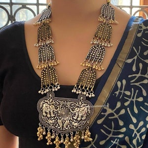 May include: A silver and gold necklace with a large pendant featuring an elephant design. The pendant is surrounded by smaller bells and the necklace is made of multiple layers of silver and gold beads.