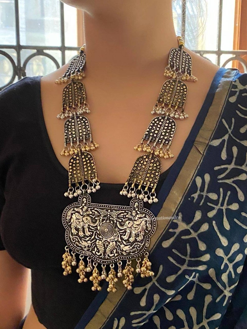 May include: A silver and gold necklace with a large pendant featuring an elephant design. The pendant is surrounded by smaller bells and the necklace is made of multiple layers of silver and gold beads.
