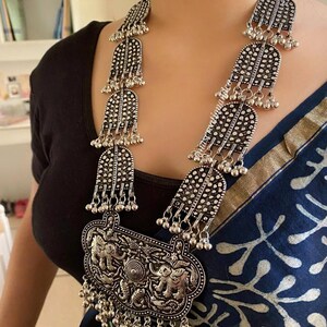 May include: A silver necklace with a large pendant featuring three elephants. The pendant is surrounded by smaller silver beads and hangs from a chain of silver beads and decorative silver pieces.