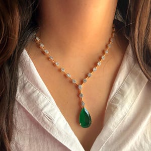 Peut inclure: Un collier en or avec un pendentif en forme de goutte d'eau en pierre verte. Le collier présente une chaîne délicate avec de petits accents étincelants. Le pendentif est d'un vert vif, et le collier est porté sur une chemise blanche.
