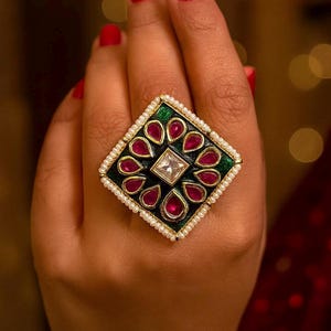 May include: A large, square-shaped ring with a black, green, and gold design. The ring features a central clear square stone, surrounded by red teardrop-shaped stones. The ring is edged with small white pearls. The ring is on a hand with red nail polish.