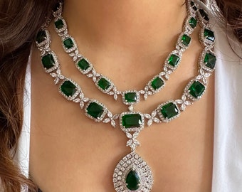 Op de afbeelding: Een zilveren ketting met een traanvormige hanger met grote smaragdgroene stenen en kleinere heldere stenen. De ketting is dubbel gelegd en heeft een delicaat bloemenmotief.