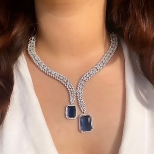 Puede incluir: Un collar deslumbrante con un diseño intrincado de pequeñas piedras transparentes. Dos gemas rectangulares de color azul oscuro cuelgan del collar. El collar se lleva sobre una blusa blanca.