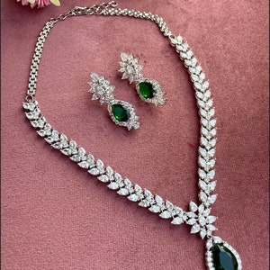 May include: A silver necklace with a teardrop-shaped pendant featuring a large green gemstone surrounded by smaller clear stones. Matching earrings with green gemstones and clear stones are also included.