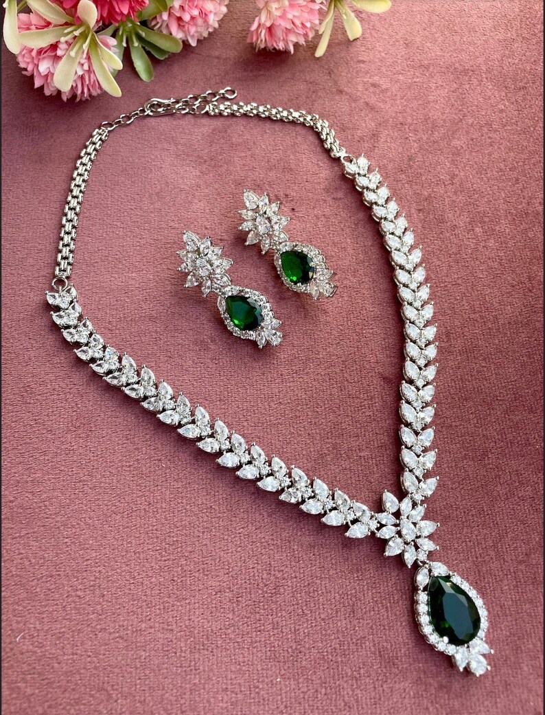 May include: A silver necklace with a teardrop-shaped pendant featuring a large green gemstone surrounded by smaller clear stones. Matching earrings with green gemstones and clear stones are also included.