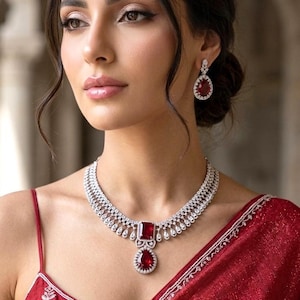 Puede incluir: Un conjunto de collar y pendientes con un gran colgante de rubí rectangular y pendientes a juego en forma de lágrima. La joyería está engastada en metal plateado o blanco. La mujer lleva un vestido rojo.
