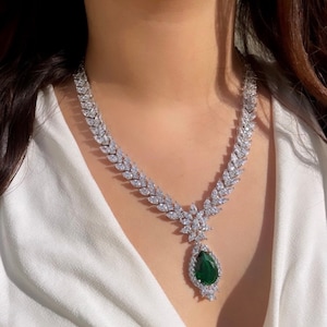 May include: A silver necklace with a teardrop-shaped green gemstone pendant surrounded by smaller clear stones. The necklace is made of a chain of clear stones in a leaf-like pattern.