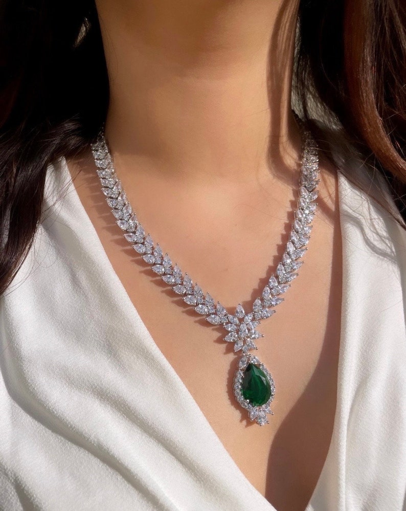May include: A silver necklace with a teardrop-shaped green gemstone pendant surrounded by smaller clear stones. The necklace is made of a chain of clear stones in a leaf-like pattern.