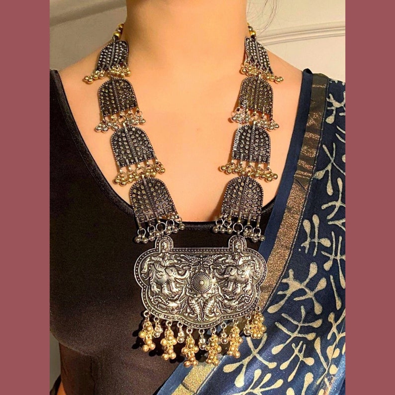 May include: A silver and gold statement necklace with a large, ornate pendant featuring a detailed design. The necklace has multiple tiers of smaller pendants and is finished with a row of gold beads.