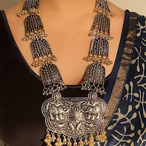 May include: A silver and gold necklace with a large pendant featuring two elephants. The pendant is ornate and has a detailed design. The necklace is made of metal beads and has a long, flowing design.
