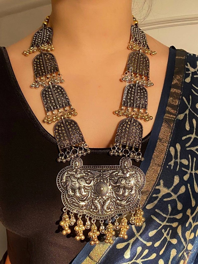 May include: A silver and gold necklace with a large pendant featuring two elephants. The pendant is ornate and has a detailed design. The necklace is made of metal beads and has a long, flowing design.