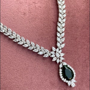 May include: A silver necklace with a teardrop-shaped green gemstone pendant surrounded by smaller clear stones. The necklace is made of a chain of clear stones in a leaf-like pattern.