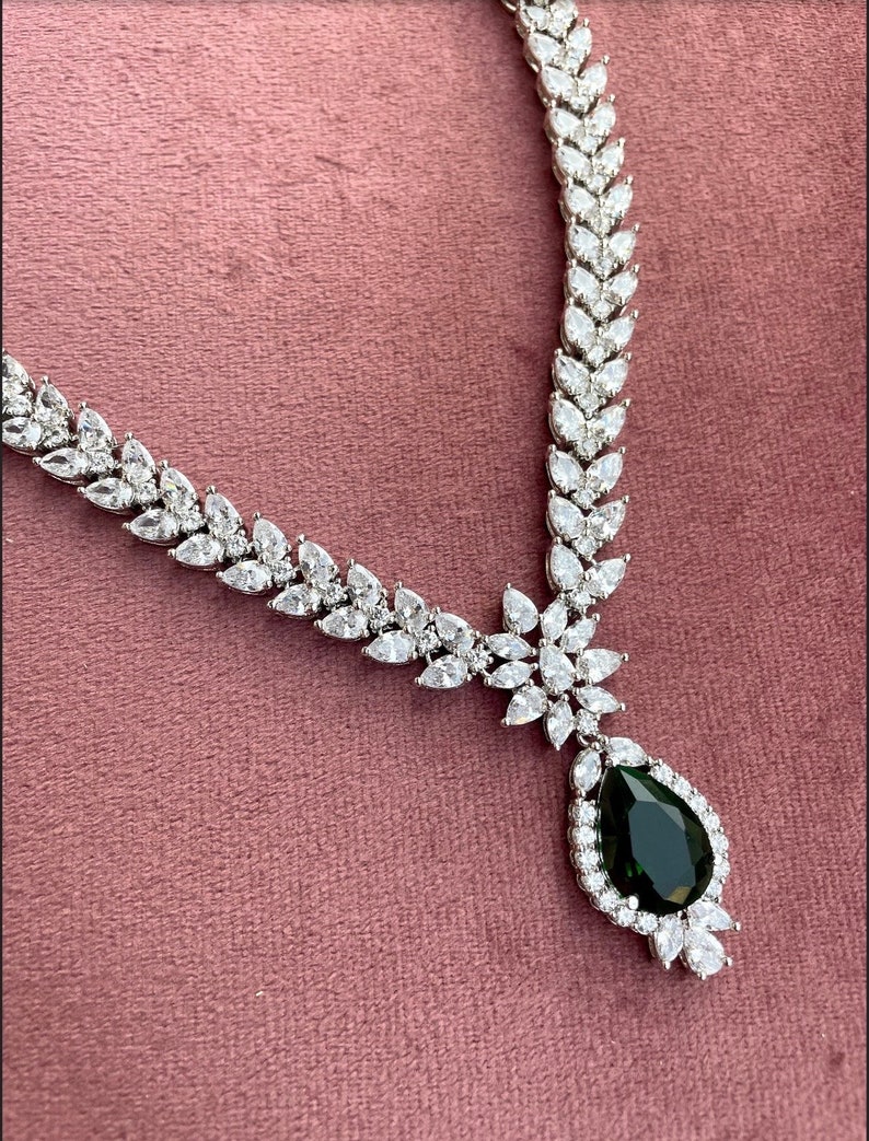 May include: A silver necklace with a teardrop-shaped green gemstone pendant surrounded by smaller clear stones. The necklace is made of a chain of clear stones in a leaf-like pattern.
