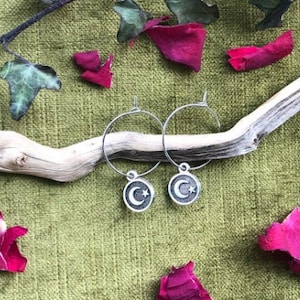 May include: A pair of silver hoop earrings with a crescent moon and star charm. The earrings are displayed on a piece of driftwood with pink rose petals scattered around.