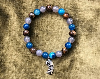Könnte beinhalten: Ein Armband mit braunen, blauen und grauen Steinen und einem silbernen Anhänger einer Seeotter. Das Armband liegt auf einem braunen Stoffhintergrund.