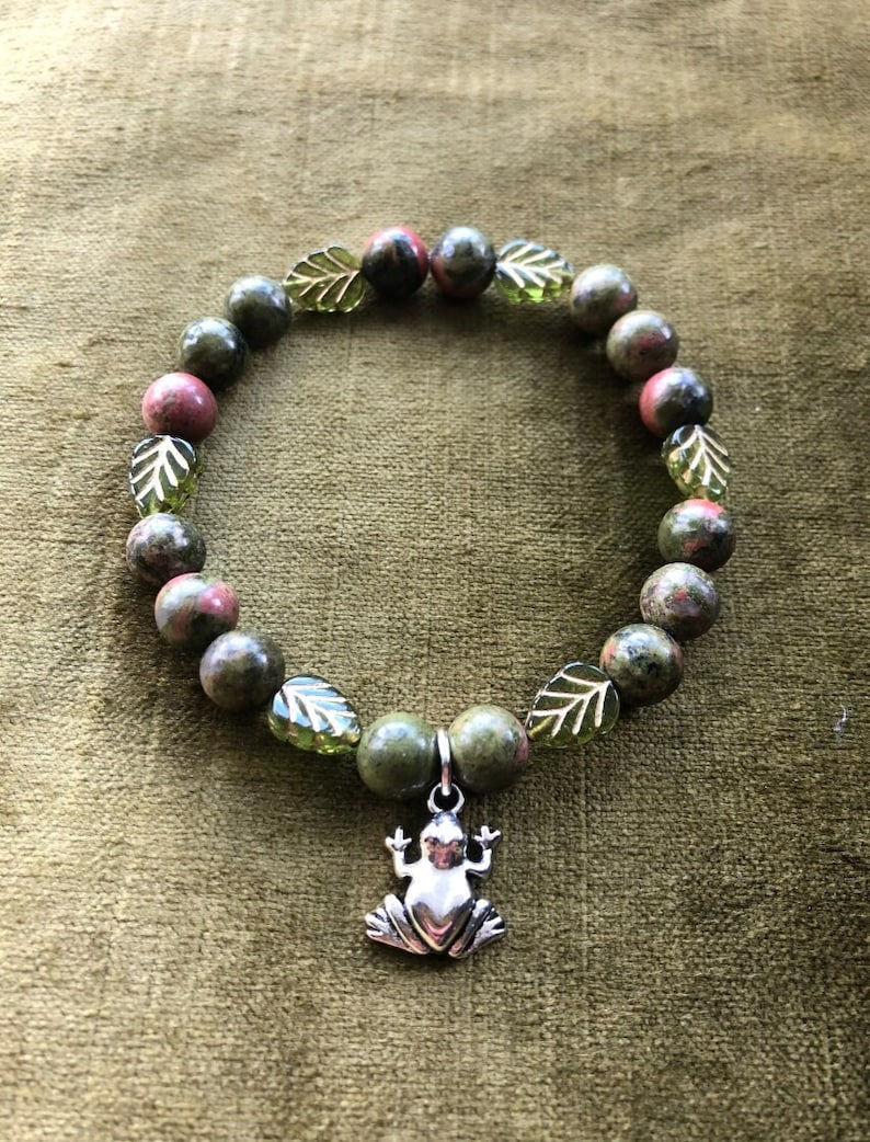 K&ouml;nnte beinhalten: Ein Armband mit gr&uuml;nen und kastanienbraunen Steinperlen, unterbrochen von gr&uuml;nen blattf&ouml;rmigen Akzenten. Ein silberner Frosch-Anh&auml;nger h&auml;ngt an dem Armband, das auf einer gr&uuml;nen Oberfl&auml;che pr&auml;sentiert wird.