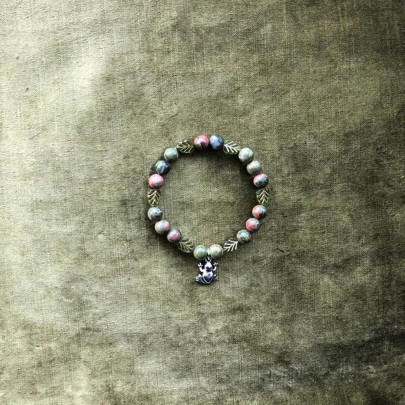 K&ouml;nnte beinhalten: Ein Armband mit gr&uuml;nen, rosa und blauen Perlen. Das Armband hat silberfarbene Blattakzente und einen Frosch-Anh&auml;nger. Pr&auml;sentiert auf einem strukturierten gr&uuml;nen Hintergrund.