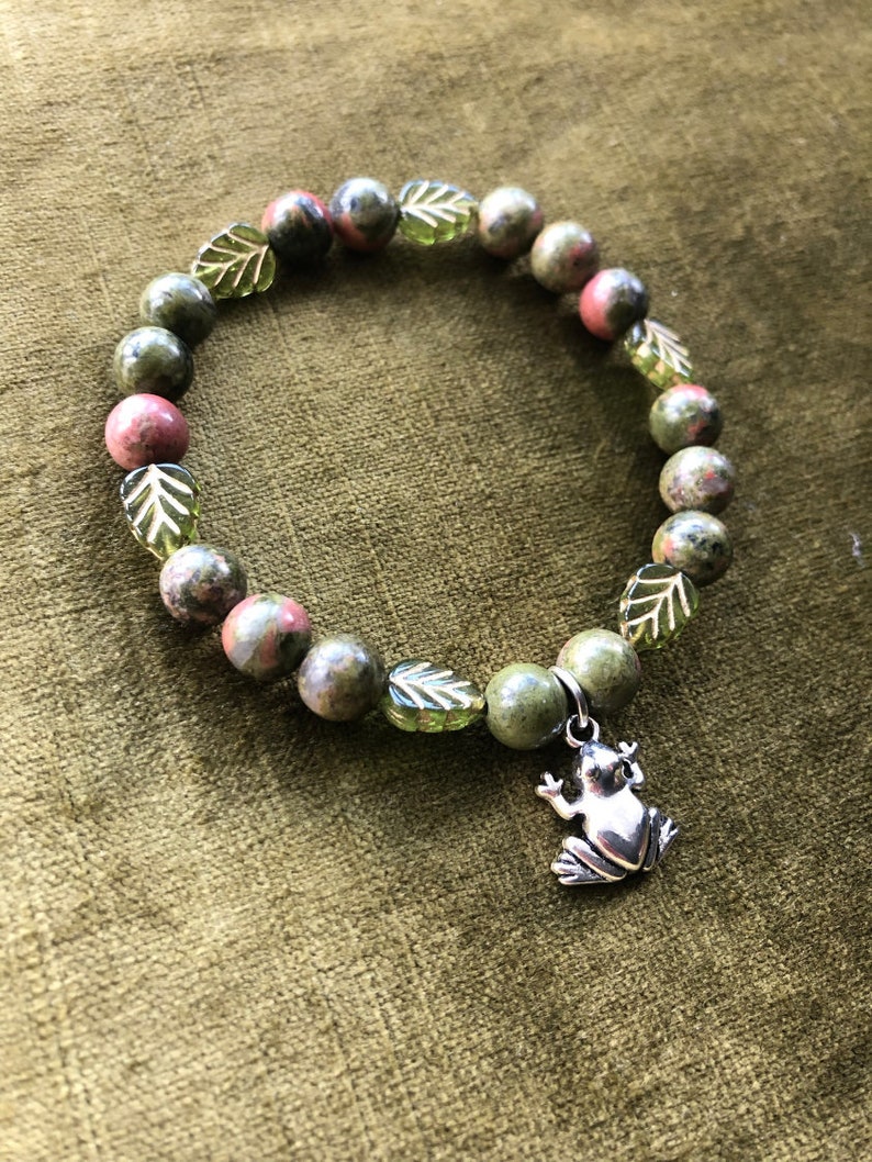 K&ouml;nnte beinhalten: Armband mit gr&uuml;nen und rosa Unakit-Steinen und gr&uuml;nen, blattf&ouml;rmigen Perlen. Ein silberner Frosch-Anh&auml;nger h&auml;ngt am Armband. Das Armband wird auf einem gr&uuml;nen Hintergrund pr&auml;sentiert. Die Perlen haben einen Durchmesser von ca. 0,8 cm.