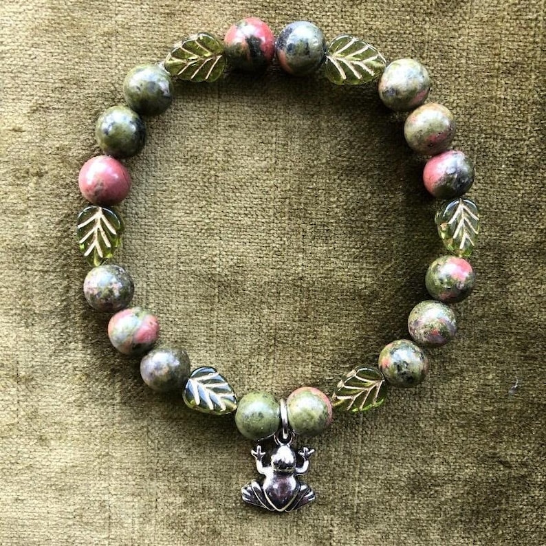 K&ouml;nnte beinhalten: Ein Armband mit gr&uuml;nen und rosa Steinperlen, die mit blattf&ouml;rmigen Glasperlen durchsetzt sind. Ein silberner Frosch-Anh&auml;nger h&auml;ngt an dem Armband. Das Armband wird auf einer strukturierten gr&uuml;nen Oberfl&auml;che gezeigt.