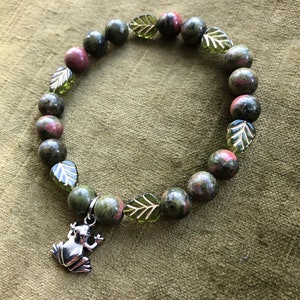 K&ouml;nnte beinhalten: Ein Armband mit gr&uuml;nen und roten Unakit-Steinen, durchsetzt mit gr&uuml;nen, blattf&ouml;rmigen Perlen. Ein silberner Frosch-Anh&auml;nger h&auml;ngt am Armband. Das Armband wird auf einem gr&uuml;nen, strukturierten Hintergrund pr&auml;sentiert.
