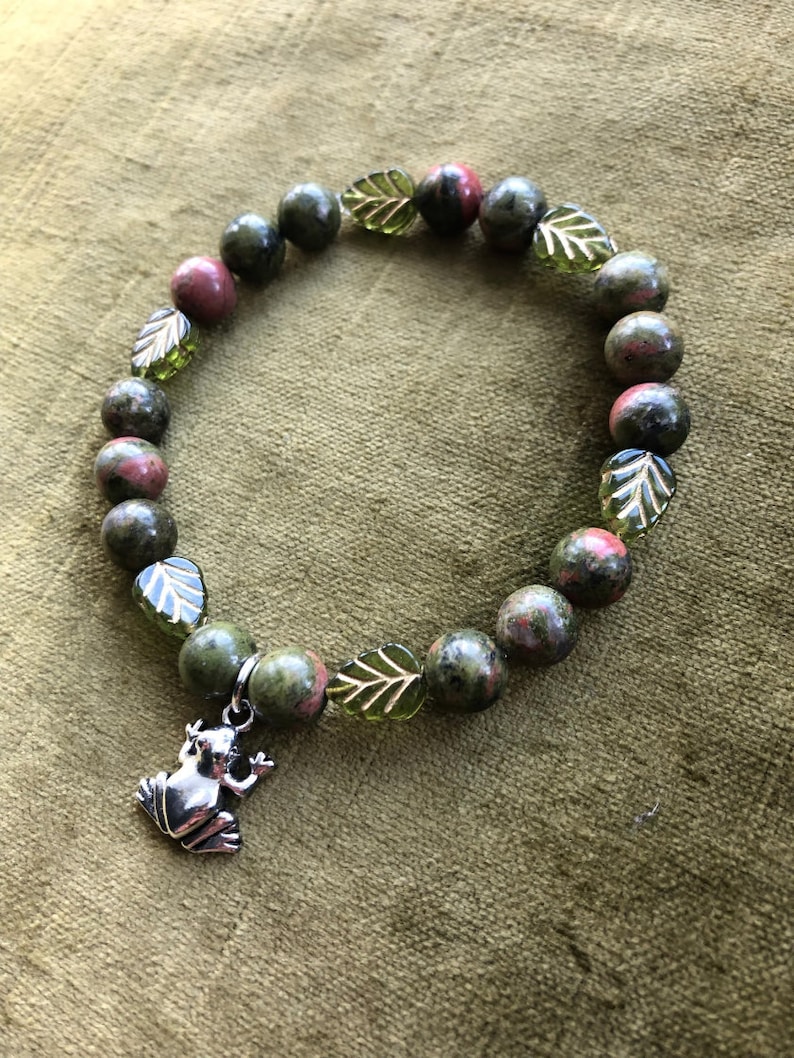K&ouml;nnte beinhalten: Ein Armband mit gr&uuml;nen und roten Unakit-Steinen, durchsetzt mit gr&uuml;nen, blattf&ouml;rmigen Perlen. Ein silberner Frosch-Anh&auml;nger h&auml;ngt am Armband. Das Armband wird auf einem gr&uuml;nen, strukturierten Hintergrund pr&auml;sentiert.