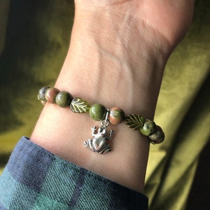 K&ouml;nnte beinhalten: Ein Armband mit gr&uuml;nen und braunen Unakit-Steinen und einem silbernen Frosch-Anh&auml;nger. Das Armband enth&auml;lt auch gr&uuml;ne, blattf&ouml;rmige Akzente. Das Armband wird an einem Handgelenk getragen.