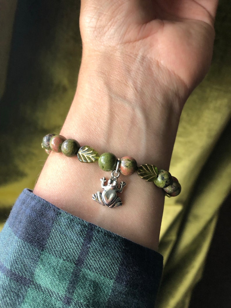 K&ouml;nnte beinhalten: Ein Armband mit gr&uuml;nen und braunen Unakit-Steinen und einem silbernen Frosch-Anh&auml;nger. Das Armband enth&auml;lt auch gr&uuml;ne, blattf&ouml;rmige Akzente. Das Armband wird an einem Handgelenk getragen.