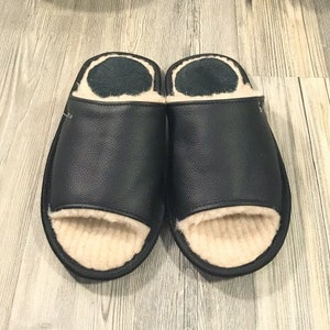 Könnte beinhalten: Ein Paar schwarze Lederschlappen mit beigefarbenem Fleecefutter. Die Hausschuhe haben ein schlichtes Design und sind perfekt für den Innenbereich.