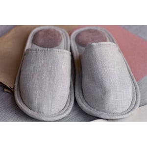Op de afbeelding: Een paar grijze linnen slippers met een bruine su&egrave;de binnenzool. De slippers zijn zacht en comfortabel, perfect om in huis te dragen.