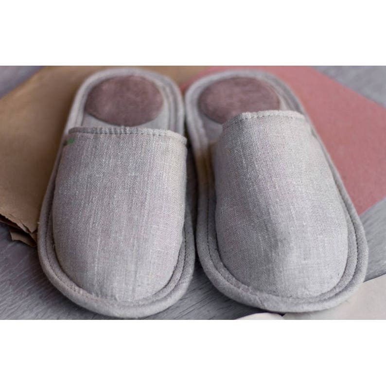 Op de afbeelding: Een paar grijze linnen slippers met een bruine su&egrave;de binnenzool. De slippers zijn zacht en comfortabel, perfect om in huis te dragen.
